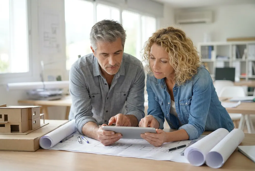 Two architect partners going over plans and a tablet at a studio table beside a model house
