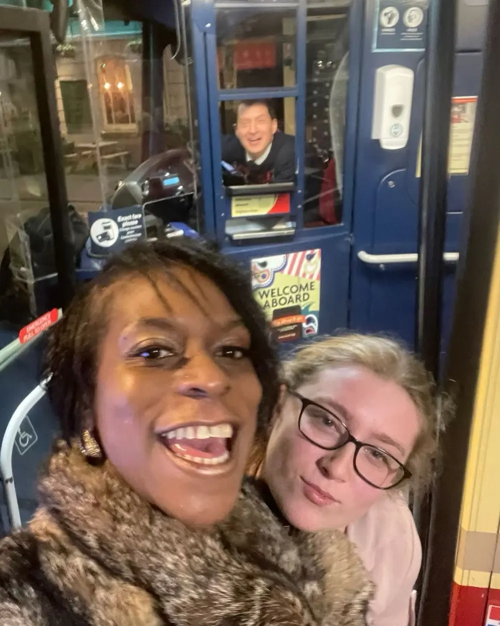 Two women grinning in a selfie aboard a vintage bus, the driver smiling behind them